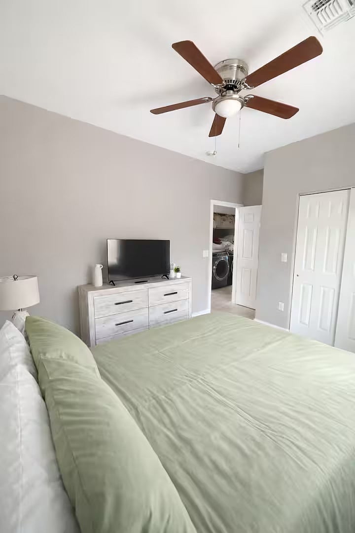 Bright and comfortable bedroom with a cozy bed, soft linens, nightstand, dresser, and TV. Natural light fills the room, and the ceiling fan keeps the space cool and comfortable. Perfect for relaxing after a long day.