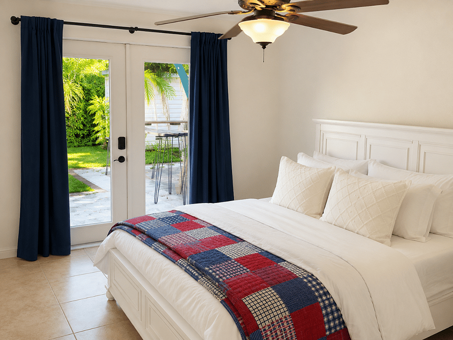 Bedroom 1 with a queen bed, simple coastal bedding, and a view of the tropical backyard.