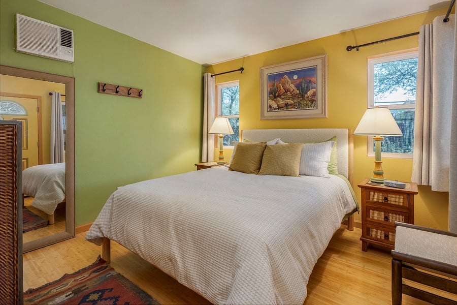 From the front door, you step directly into a cheerful bedroom. A plush queen bed sits beneath an original hand-painted desert landscape, flanked by bamboo nightstands and matching mid-century lamps; A bedside digital clock serves as a smartphone charger.  Warm bamboo flooring and a colorful vintage Turkish kilim rug ground the room.