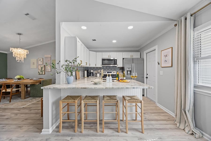 A refined coastal-modern kitchen with premium finishes, a spacious island, and elegant seating for four.  White cabinetry, gold hardware, and warm woven stools create an elevated, boutique-stay feel, perfect for relaxed mornings or stylish evenings in.