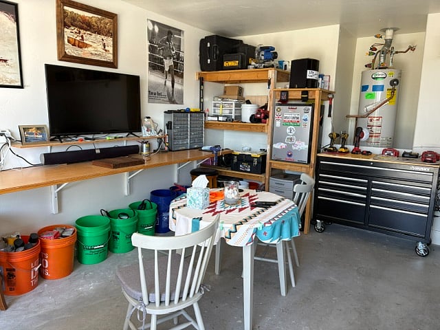 Garage space with table, 2 chairs, mini fridge, and TV with a soundbar and subwoofer. 