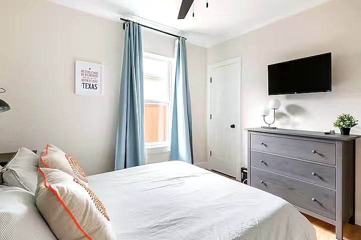 Bedroom 2 offers a comfortable bed with soft linens, a wall-mounted smart TV, dresser storage, and room-darkening curtains. Clean, cozy, and thoughtfully styled for a restful stay.