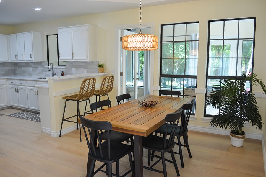 Fully equipped kitchen with white shaker cabinets, marble tile backsplash, and breakfast bar seating for 2. Dining table seats 6. Door opens to sunroom and backyard beyond. Steel-frame windows with lush tree views throughout.