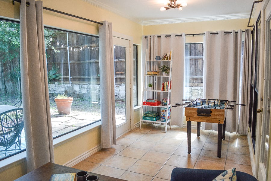 Sunroom game room with foosball table, bookshelf stocked with novels and board games, and large windows overlooking the backyard patio with caf string lights. Door opens directly to outdoor seating area.