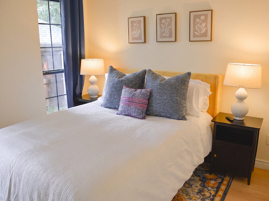 Second bedroom with queen bed, mustard velvet headboard, hotel-quality linens, dual nightstands, botanical art, and vibrant Persian-style rug. Closet and smart TV included. Steel-frame window with natural light.