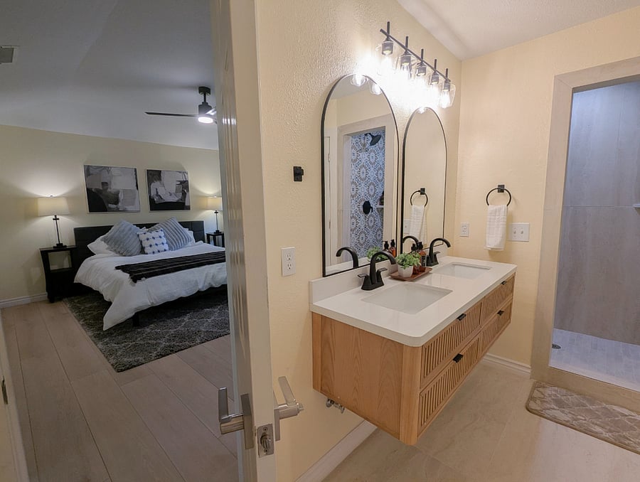 Primary suite with direct ensuite access king bedroom and spa bathroom in one view. Double vanity with floating wood cabinet, quartz countertops, dual arch mirrors, black matte fixtures, and walk-in shower with Moroccan tile.