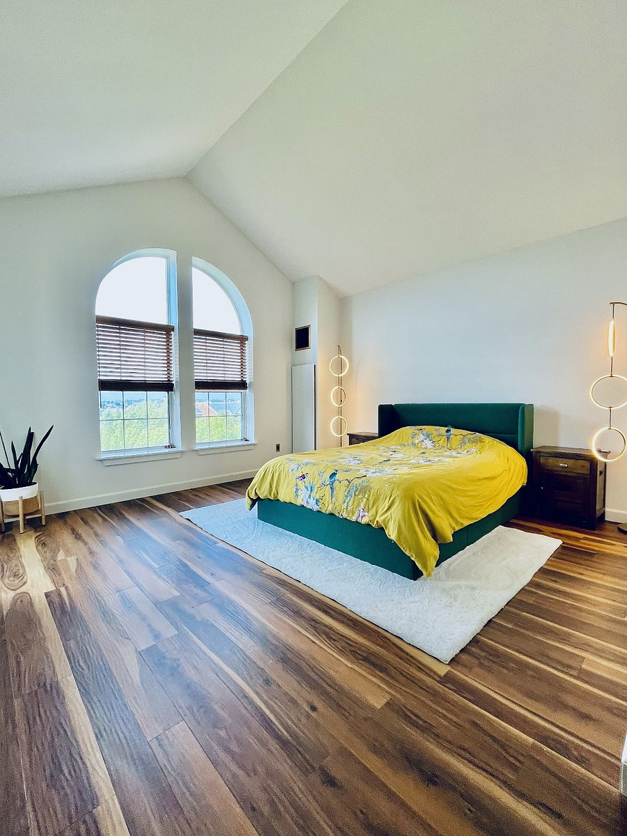 Expansive primary bedroom with cathedral ceilings, warm wood flooring, an upholstered bed with ample under-bed storage, and dramatic arched windows.