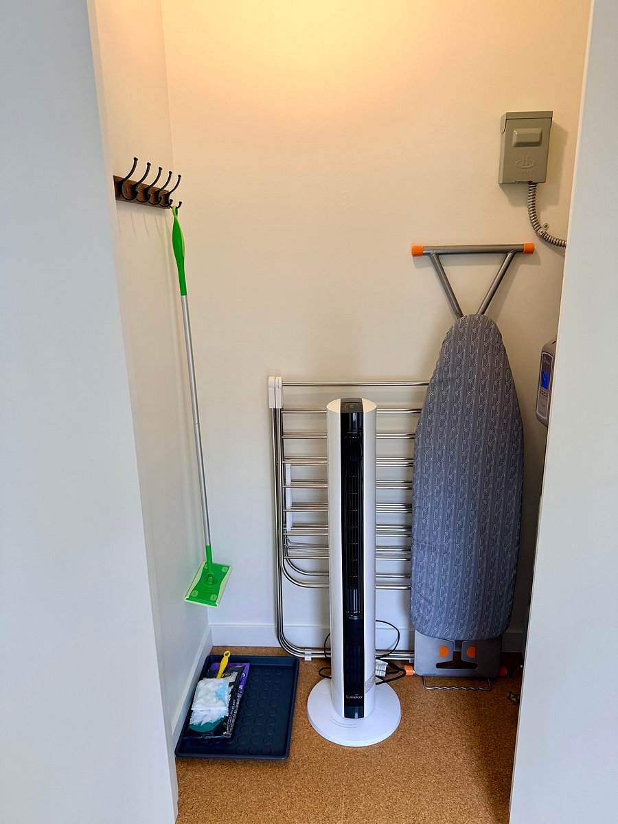 Inside entryway closet, with coat rack, shoe tray, ironing board, clothes drying rack, tower stand fan & heater, and cleaning supplies.