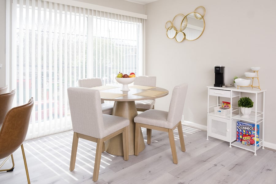 A sun-drenched and welcoming dining area featuring an elegant round table with cream-colored chairs and a charming coffee bar.