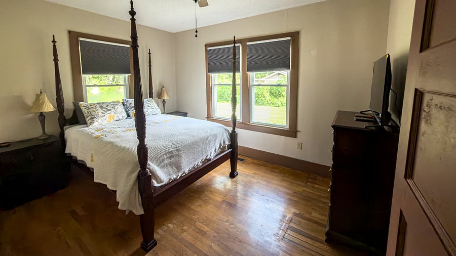 Another angle on the master four-poster queen, dual nightstands with USB charging, smart TV, and blackout shades on every window. The hardwood floors, trim, and doors are all original to the house.