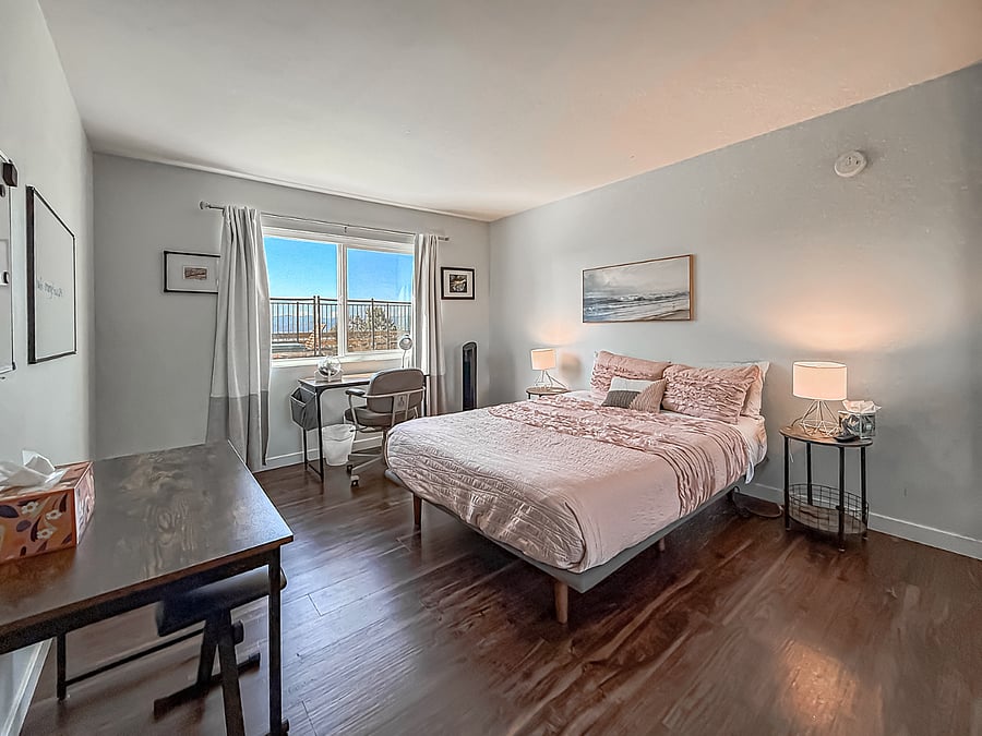 Spacious furnished bedroom with wood flooring, floor-to-ceiling window, private balcony, updated decor, and ample space for sitting area and storage.