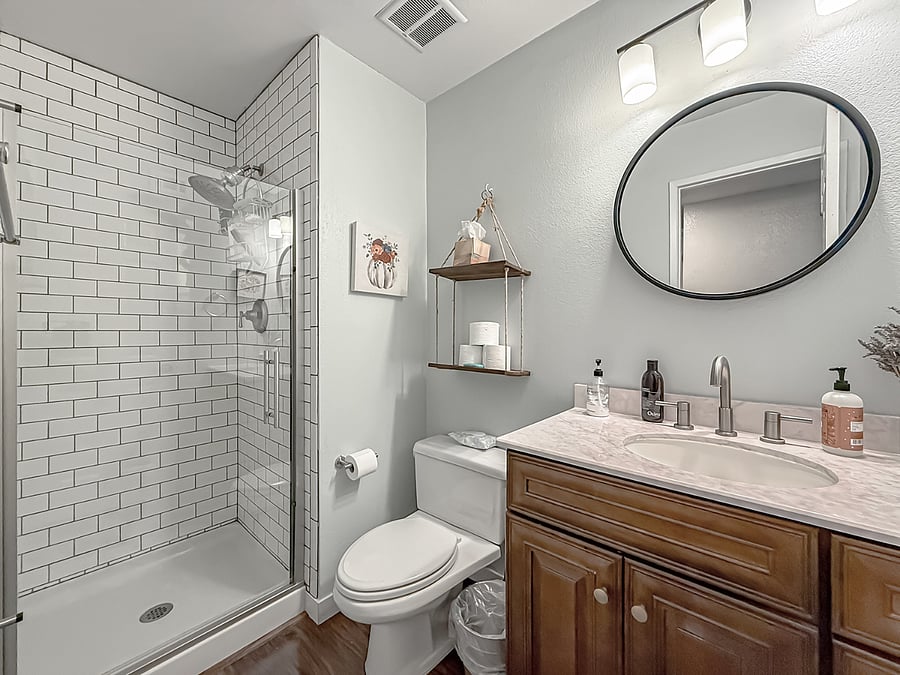 Fully furnished full bath with marble vanity, wood cabinetry, round mirror, glass-enclosed shower with subway tile, and modern accessories.