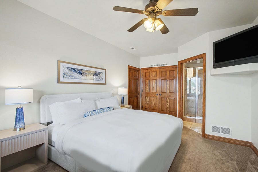 Spacious primary bedroom featuring a new comfortable king-size mattress, new bedding and furniture, a wall-mounted TV, ceiling fan, generous closet space, and direct access to the separate shower and vanity area.