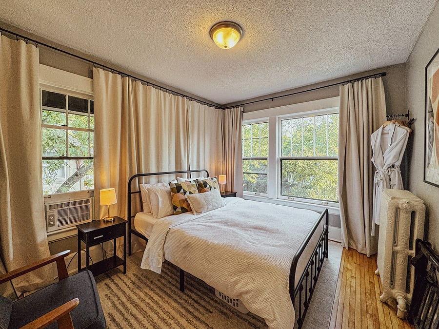Spacious queen bedroom with organic linens, beautiful natural light, blackout window coverings, closet storage, a cozy bathrobe, and easy bedside phone charging.
