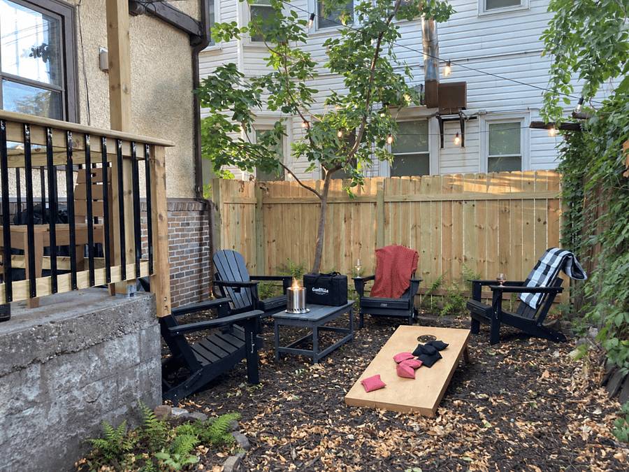 Unwind in the backyard beneath the bistro lights with Adirondack chairs, a tabletop pellet firepit, cornhole, and lawn games. The yard is partially fenced but not fully enclosed.