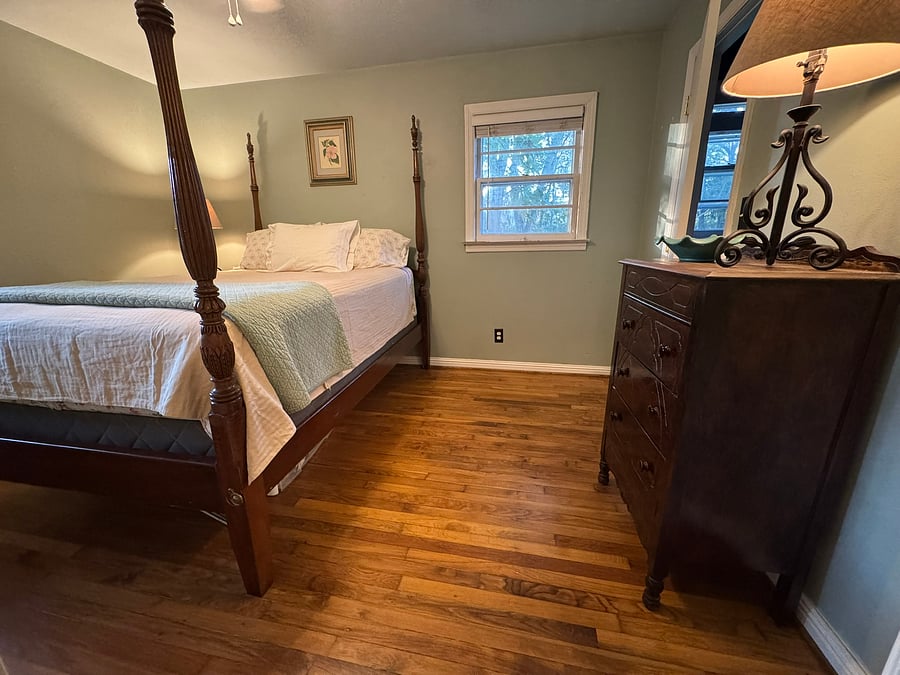 The serene primary bedroom is the perfect place to relax, unwind, and snuggle up. Includes a four-poster bed, lamps, dresser, and closet. Half bath is through open door on the right. Overlooks the side yard and backyard.