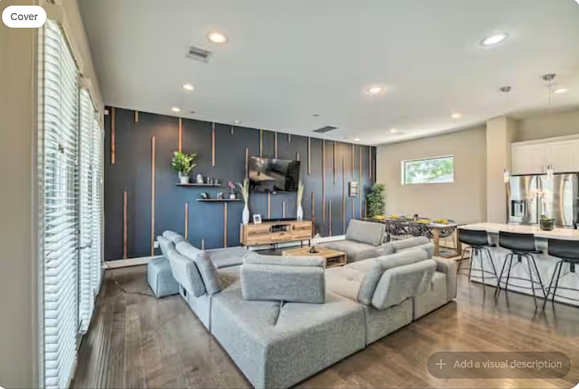 Bright, modern living room with a large gray sectional, warm wood floors & statement TV wall.
Large windows add natural light.
Open layout flows into the dining area & white kitchen with island seatinggreat for relaxing & gathering.