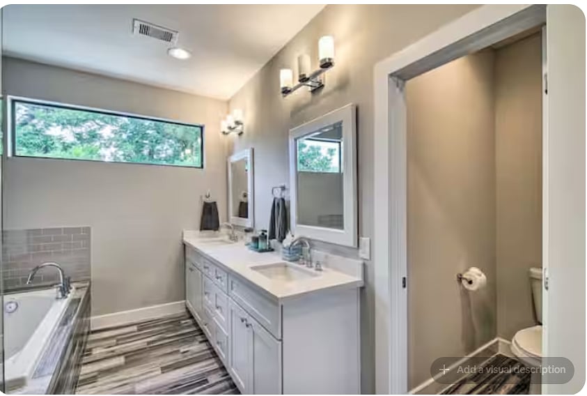 Spacious en-suite bathroom with a long dual-sink vanity, large mirrors & modern lighting.
Features wood-look tile flooring, a soaking tub and separate toilet area.
Wide window brings in natural light while maintaining privacy.