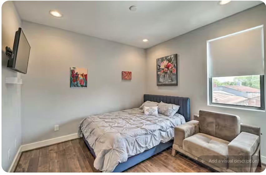 Comfortable bedroom with an upholstered bed, warm wood floors & cozy sitting chair.
Wall-mounted TV adds convenience for relaxing.
Colorful artwork and recessed lighting add character, while a large window provides natural light.
