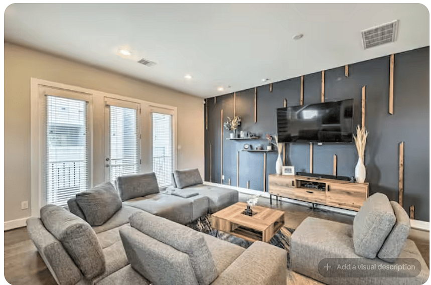 Inviting living room with a spacious gray sectional, warm wood floors & modern coffee table.
Large TV sits against a striking dark accent wall with natural wood detailing.
Glass doors bring in natural light and provide outdoor access.