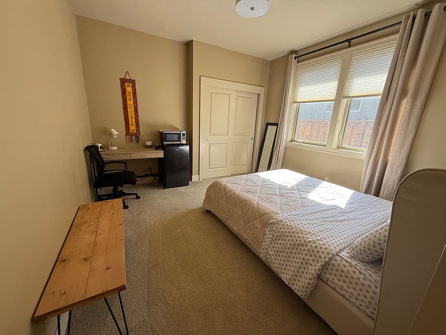 The room includes a comfortable bed with an upholstered headboard, bedding, a dedicated desk and workspace, a large closet, mini refrigerator, and microwave. There is plenty of open floor space, making the room feel comfortable for an extend day.