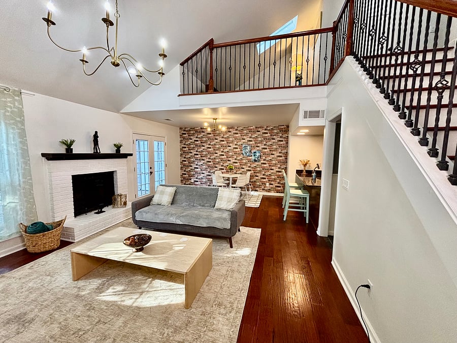Spacious high-ceiling living room featuring natural light, hardwood floors, cozy fireplace, and 55" Smart TV.