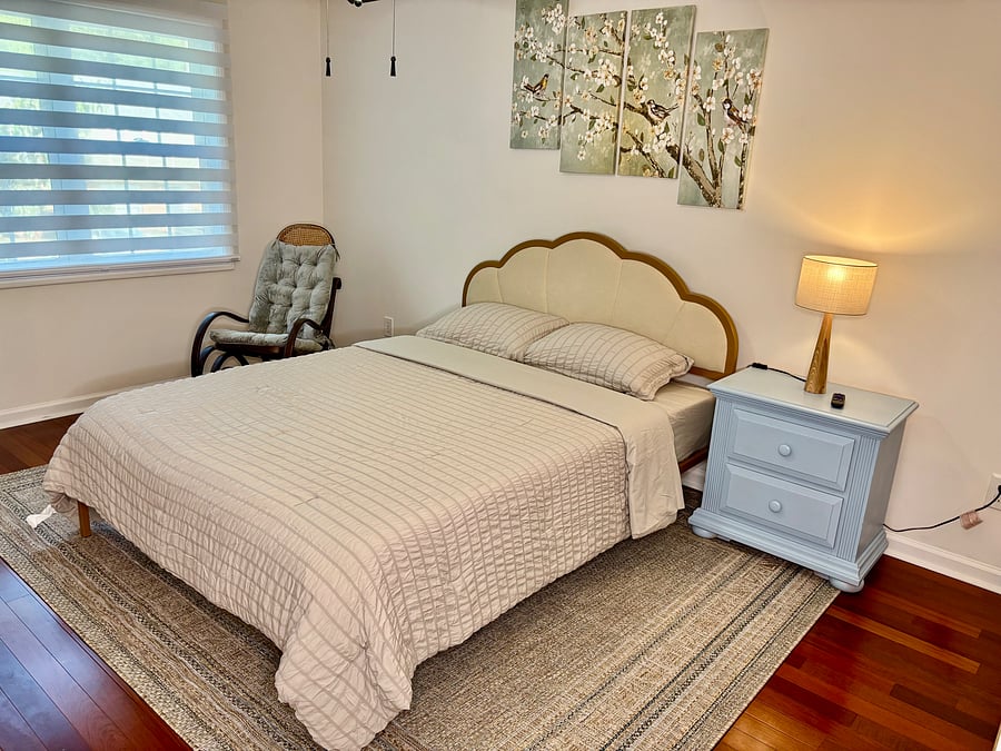 Comfortable bedroom with cozy queen bed, 32" smart tv, makeup table with mirror ,bedside table, rocking chair, and zebra shades for light control.