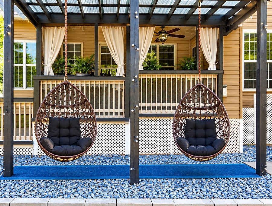 Twin woven hanging chairs with plush black cushions offer an inviting place to unwind beneath the pergola. Airy curtains and greenery on the adjoining deck create a relaxed, resort-inspired backdrop for quiet conversation or an unhurried afternoon.