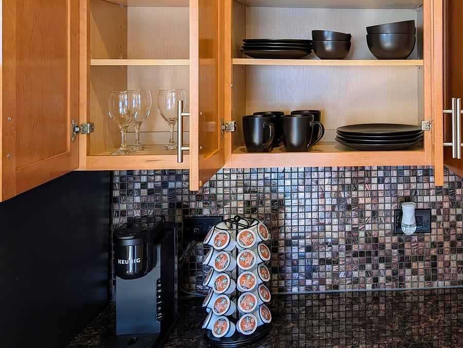 Kitchen: Fully equipped upper cabinets featuring dinnerware plates, bowls, coffee mugs, and wine glasses, paired with a Keurig coffee station and pod carousel.