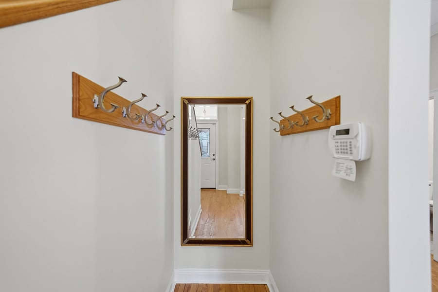 Front Entryway: Welcoming front entryway featuring dual wall-mounted coat racks with brass hooks, a full-length mirror, warm hardwood floors, and a dedicated security keypad panel.