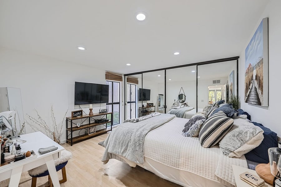 The spacious bedroom features a plush bed with layered linens, abundant pillows, a large mirrored wall, and a wall-mounted TV for relaxing in comfort.