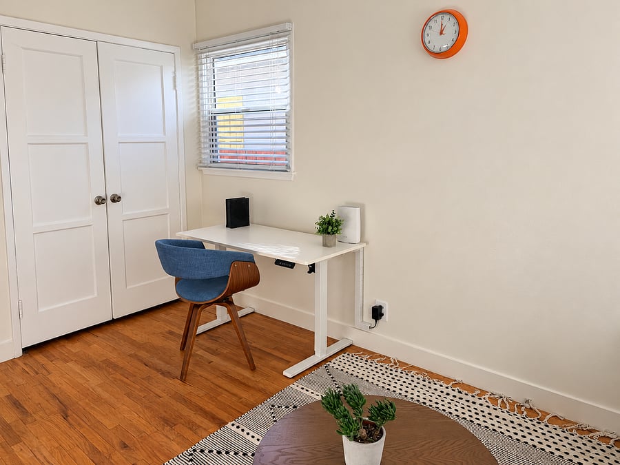 Dedicated home office with an electric height-adjustable standing desk, comfortable upholstered chair, closet storage, and natural light. Ideal for remote work.