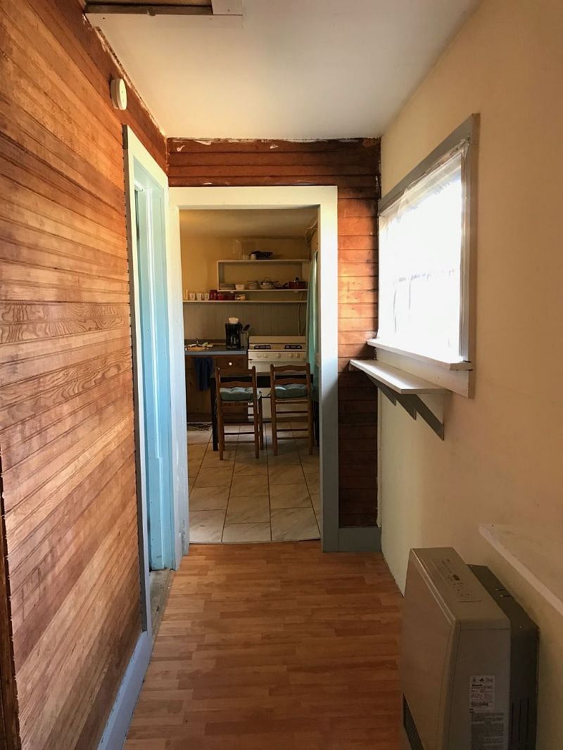 Many aspects of the cottage are original, including the tongue and groove walls of the bedroom and hallway to the bedroom. All utilities and conveniences are updated and modernized. In this photo, you are looking from the bedroom, past the bathroom door to the left and into the kitchen. There is a large and comfortable shower as well as the sink, mirror and toilet in the bathroom.