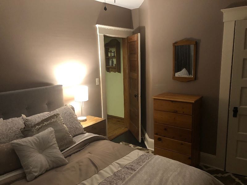 The bedroom is cozy and comfortable with a queen bed, side table with clock radio and lamp, a chest of drawers with a vanity mirror, and closet.