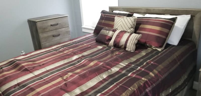 The Brown Room - Features (1) soft but firm Queen Bed with plush pillows, a dresser, night stand & lamp with colored smart bulb controlled by Google Assistant.