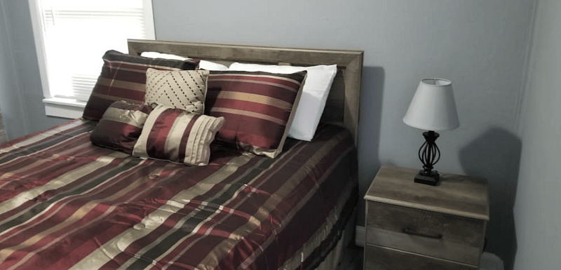 The Brown Room - Features (1) soft but firm Queen Bed with plush pillows, a dresser, night stand & lamp with colored smart bulb controlled by Google Assistant.
