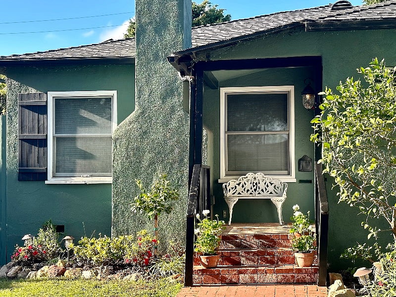 A white wrought iron bench, potted geraniums and seasonal wreath on the front door of the covered porch entrance, marks the main house residence entry.