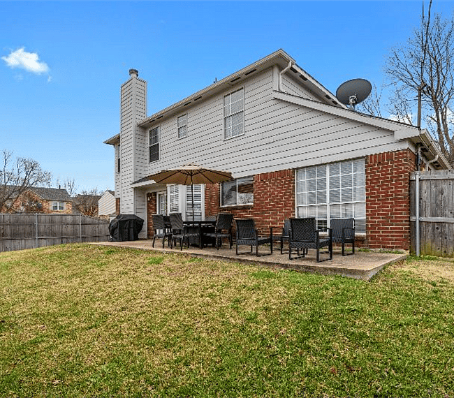 Large grassy fenced in backyard with open patio set up with dining table for 6 and sitting area.