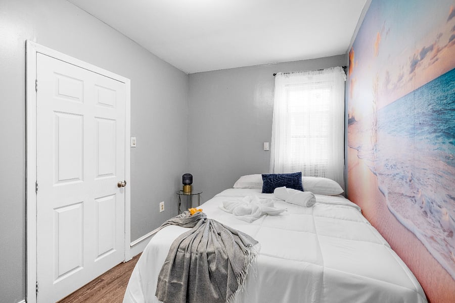 A queen-sized bed in a white bedroom accented by modern beach-themed wallpaper evokes a serene coastal vibe, blending clean, minimalist design with soothing ocean-inspired elements.