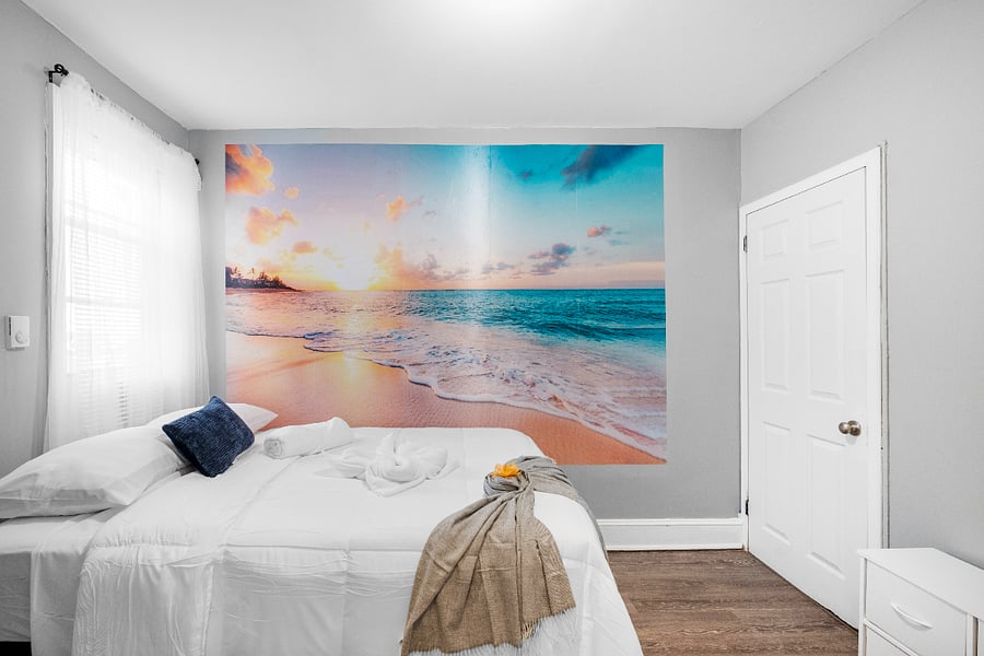 A queen-sized bed in a white bedroom accented by modern beach-themed wallpaper evokes a serene coastal vibe, blending clean, minimalist design with soothing ocean-inspired elements.