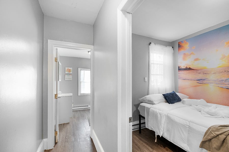 A queen-sized bed in a white bedroom accented by modern beach-themed wallpaper evokes a serene coastal vibe, blending clean, minimalist design with soothing ocean-inspired elements.