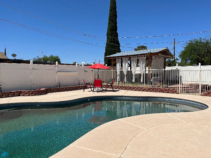 Backyard oasis has large, in-ground pool, multiple patio tables, and decorated and light pergola.