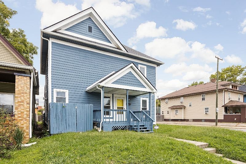Side angle view of a beautifully updated blue two-story home featuring a cozy covered porch, white trim accents, and a fenced backyard.