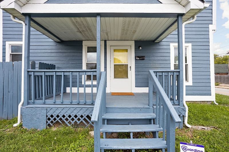 Inviting front view of a cozy blue house with white trim, featuring a covered porch, wooden railings, and a small staircase leading up from a well-kept lawn.