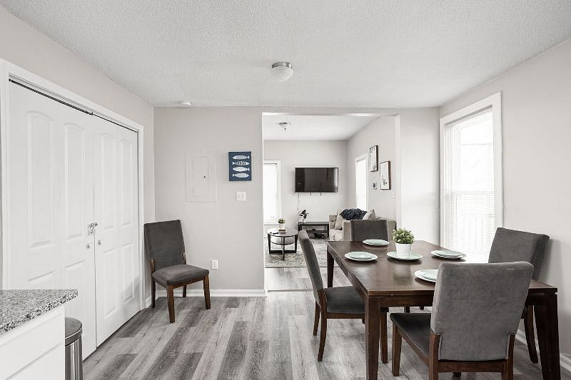 Bright and inviting dining area with a sleek wooden table set for four, stylish gray upholstered chairs, and a seamless view into the cozy, modern living room beyond.