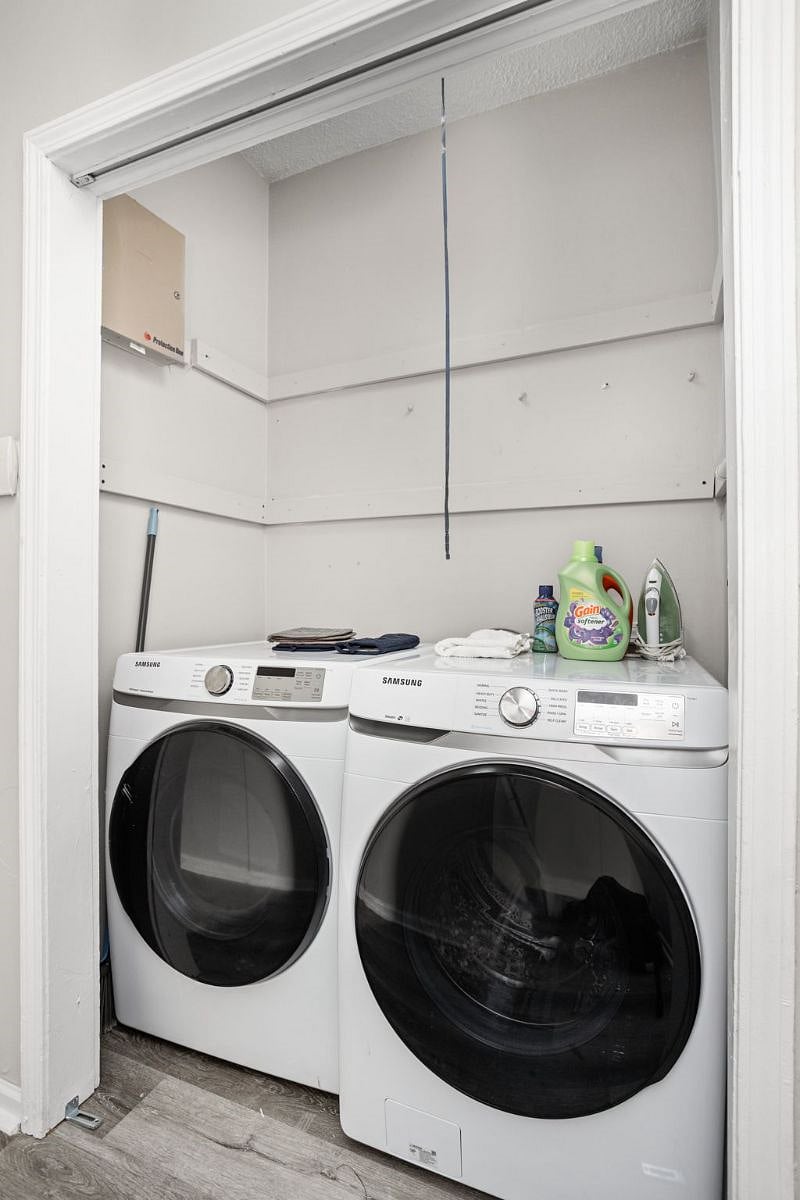 Compact laundry nook featuring a modern Samsung washer and dryer set, neatly stocked with laundry essentials including detergent, fabric softener, and an iron.