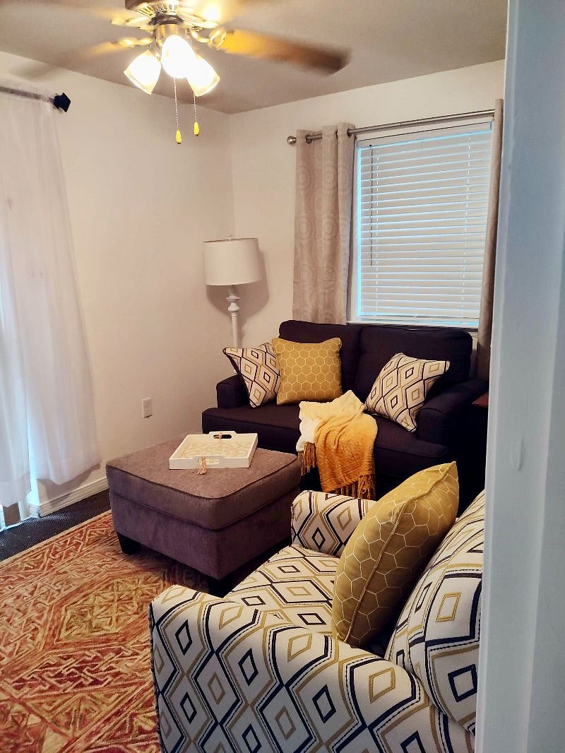 Bright and airy living room designed for comfort and relaxation. Features include a cozy sofa, club chair, ottoman, and a floor lamp for ambient lighting. A ceiling fan keeps the space cool and breezy, while the smart TV makes it easy to stream your favorite shows during downtime.W
