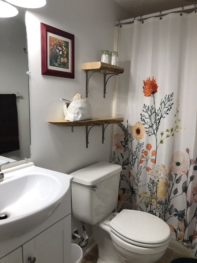 brightly lit bathroom with dressing mirror, floral shower curtain, fabric curtain liner, two towel racks, and over the door hook