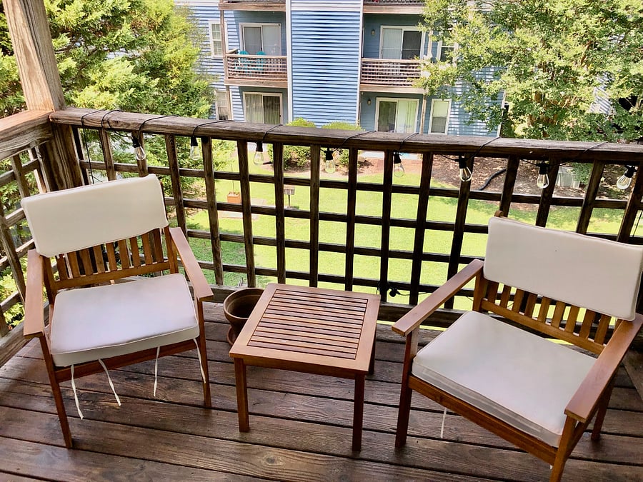 A cozy wooden balcony with two chairs and a small table, string lights strung across a railing, a grassy courtyard in the background.