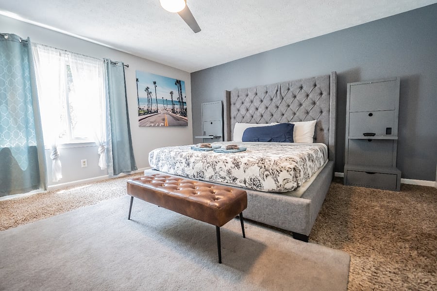 Elegant bedroom with a gray tufted headboard, soft floral bedding, a leather bench, and a large sunlit window.